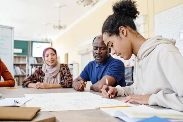 Young Black immigrant sitting at table writing something on handmade educational English language poster during lesson