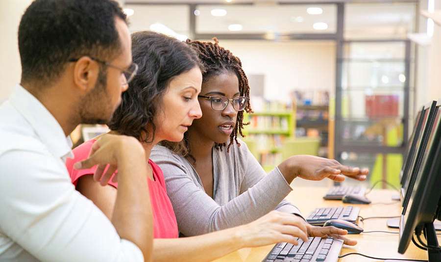 Instructor explaining corporate software specific to trainees in computer class. Man and women sitting at table, using desktop, pointing at monitor and talking. Training concept